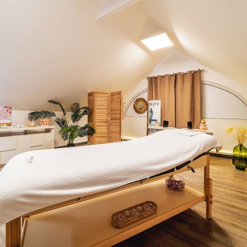 Ein gemütlicher Wellnessraum mit einem Massagetisch, der mit einem weißen Handtuch bedeckt ist, umgeben von Zimmerpflanzen und warmem, weichem Licht unter einer schrägen Decke.