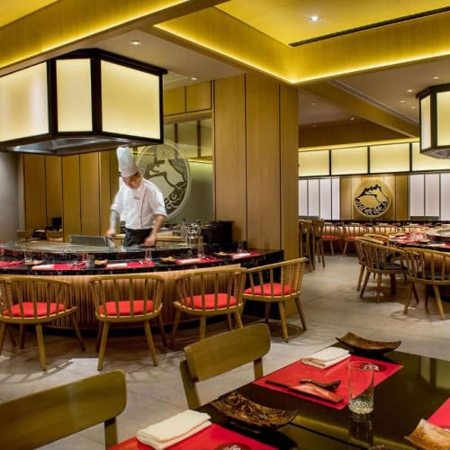 Elegantes japanisches Restaurant mit einem Sushi-Thekenbereich, einem Koch in Uniform, der Sushi zubereitet, und Tischen mit roten Tischsets und schwarzen Tellern.