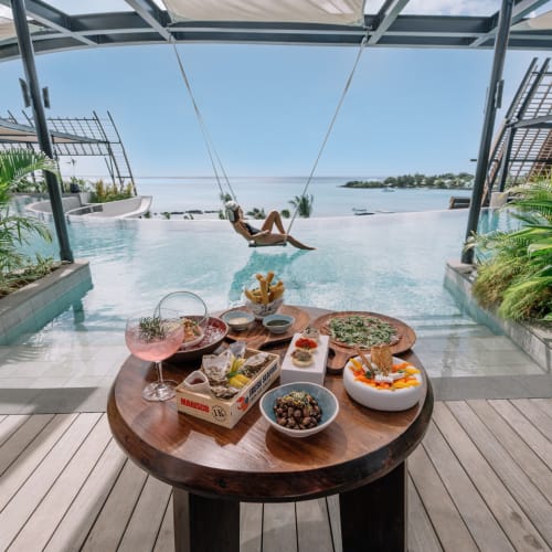 Ein Tisch mit Essen und Getränken auf einem Dachpool-Deck, im Hintergrund schwingende Person mit Blick auf den Ozean.