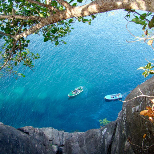 Blick von einer felsigen Klippe auf zwei kleine Boote in klarem türkisfarbenem Wasser, eingerahmt von Baumzweigen.