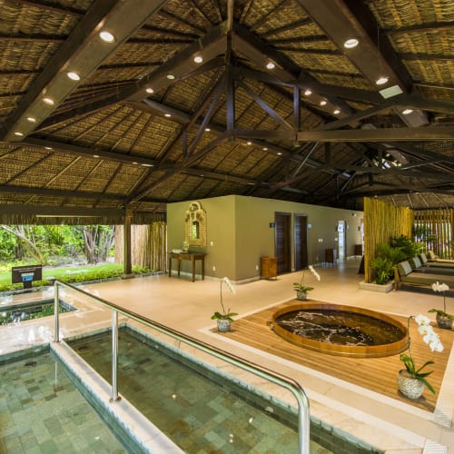 Innenbereich eines Spa-Bereichs mit Hot Tub, Lounge-Sitzgelegenheiten und üppigem Grün durch Fenster sichtbar.