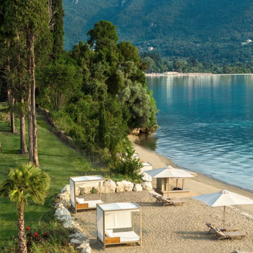 A serene beach at Ikos Odisia with sandy shore, sun loungers under white umbrellas, and lush green trees along the edge, overlooking calm blue waters and distant mountains.