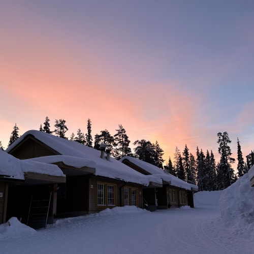 Schneebedeckte Holzhäuser entlang eines verschneiten Weges, umgeben von hohen Kiefern, unter einem farbenfrohen Sonnenuntergangshimmel in einer Winterlandschaft.