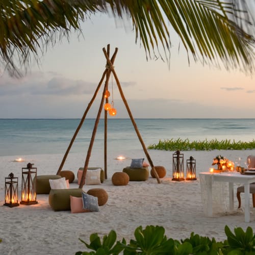Eine romantische Strandgestaltung bei Sonnenuntergang mit Esstisch, Stühlen, Laternen und Sitzgelegenheiten unter einer Palme mit Blick auf den Ozean.