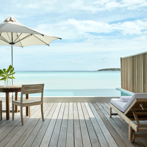 A relaxing deck scene at a Maldivian water villa with a table, chairs, an umbrella, and a lounge chair overlooking the ocean.