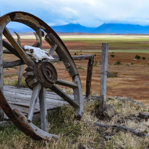 Ein alter, rostiger Wagenrad lehnt an einer Holzkonstruktion in einer offenen Ebenenlandschaft mit Bergen im Hintergrund.