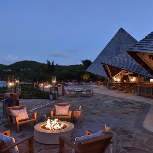 Eine Terrasse im Freien mit Holzmöbeln und einem zentralen Feuerkorb, mit Meerblick bei Dämmerung, im Hintergrund eine moderne Pavillon-Struktur.