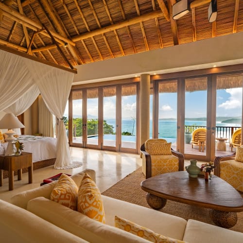 Luxuriöses Schlafzimmer und Sitzbereich mit Himmelbett und gelb gemusterten Sesseln, mit Blick auf einen Balkon und den Ozean unter einem Strohdach.