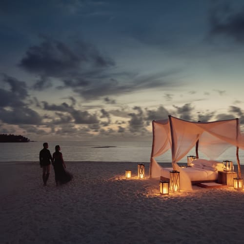 Eine romantische Strandkulisse bei Sonnenuntergang mit einem Himmelbett, umgeben von Laternen, und einem Paar, das am Ufer spazieren geht.