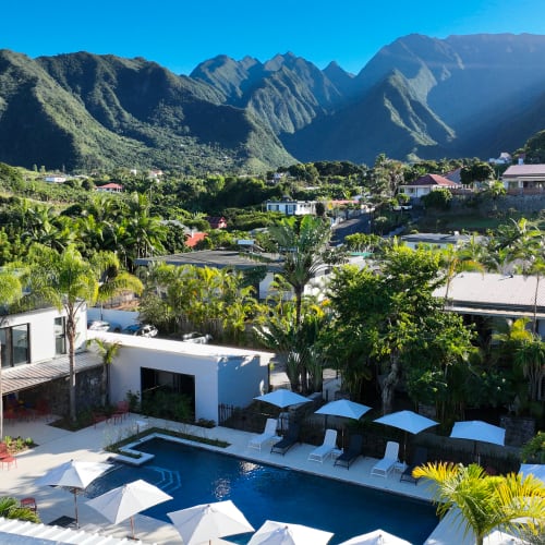 Ferienanlage mit einem Schwimmbecken, umgeben von Liegestühlen und Sonnenschirmen, eingebettet in ein grünes Tal mit dramatischen Berggipfeln im Hintergrund unter klarem blauem Himmel.