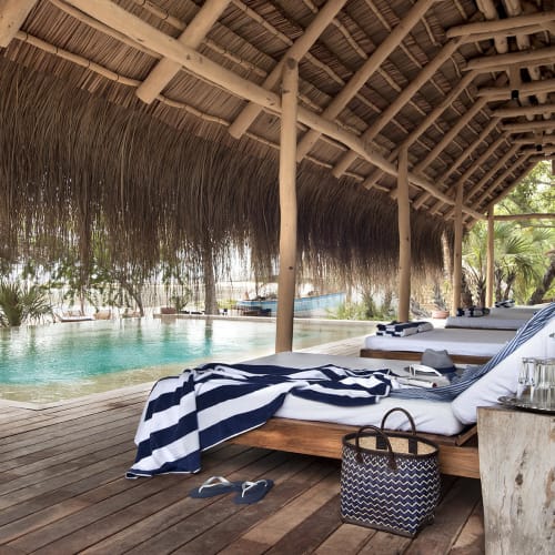Luxurious loungers with towels and drinks next to a swimming pool under a thatched roof on a tropical island.