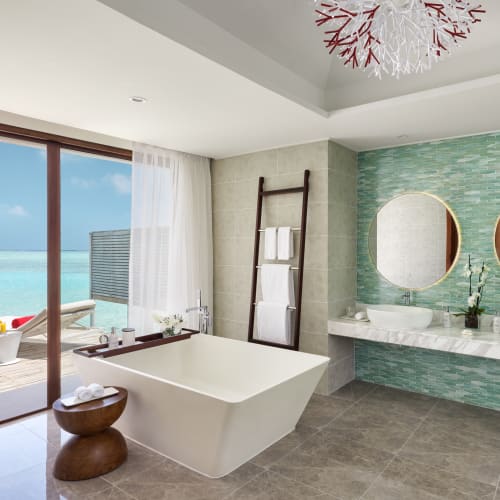 Modernes Badezimmer in einer Überwasser-Villa mit freistehender Badewanne, zwei Waschbecken mit runden Spiegeln und Blick auf ein Deck mit klarem blauem Meerwasser.