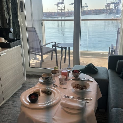 Ein Frühstückstisch in einer Suite mit Tellern voller Essen und Getränken, neben einer Glastür auf einen Balkon mit Blick auf einen Hafen mit Kränen und Wasser.