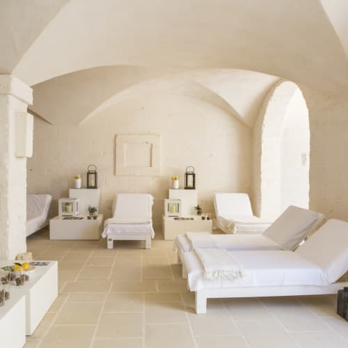A minimalist spa interior with white lounge chairs, small tables with candles, and a ladder holding towels in a bright room with arched ceilings.