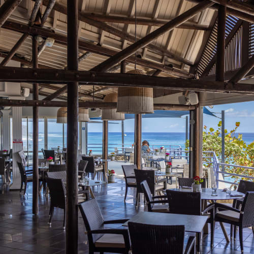 Außenbereich eines Strandrestaurants mit Tischen und Stühlen unter einem Holzpergola mit Blick auf den Ozean an einem sonnigen Tag.