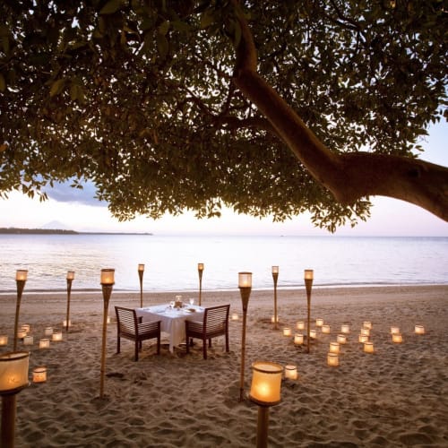 Ein romantisches Abendessen am Strand mit brennenden Kerzen und einem Tisch mit zwei Stühlen unter einem großen Baum am Strand bei Sonnenuntergang.