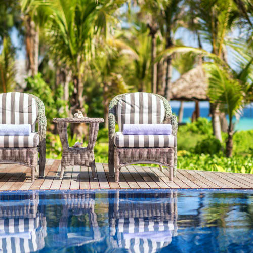 Zwei gestreifte Liegestühle mit einem kleinen Tisch dazwischen stehen neben einem Swimmingpool, im Hintergrund ein tropischer Strand und Palmen.