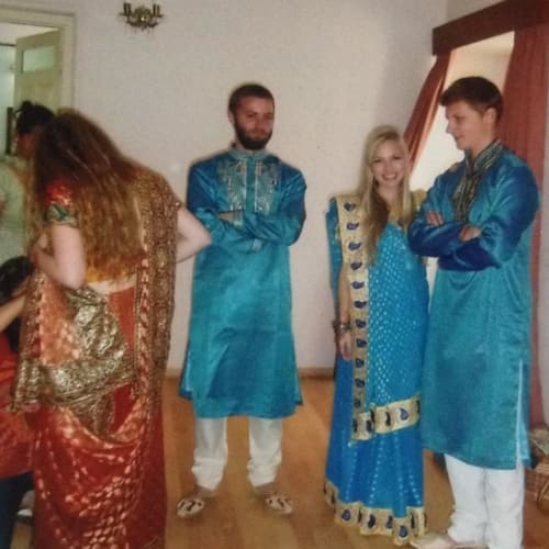 Eine Gruppe von Menschen in traditioneller indischer Kleidung, die drinnen auf einem Holzboden stehen. Zwei Männer und eine Frau tragen blaue Festkleidung, während eine weitere Frau einen roten und goldenen Sari trägt.