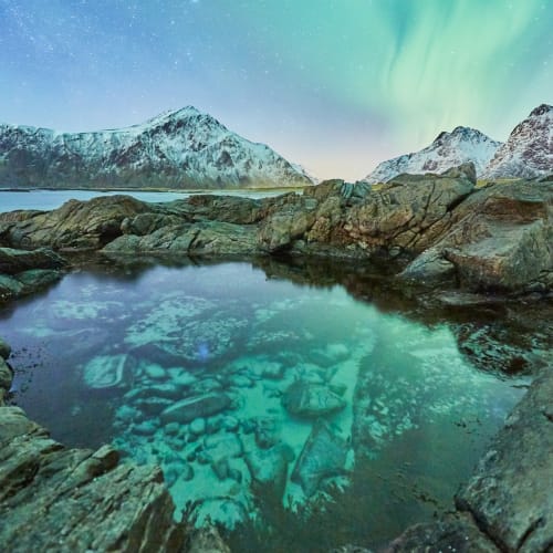 Nordlichter erleuchten den Nachthimmel über schneebedeckten Berge und eine felsige Küste mit natürlichem Thermalbecken.