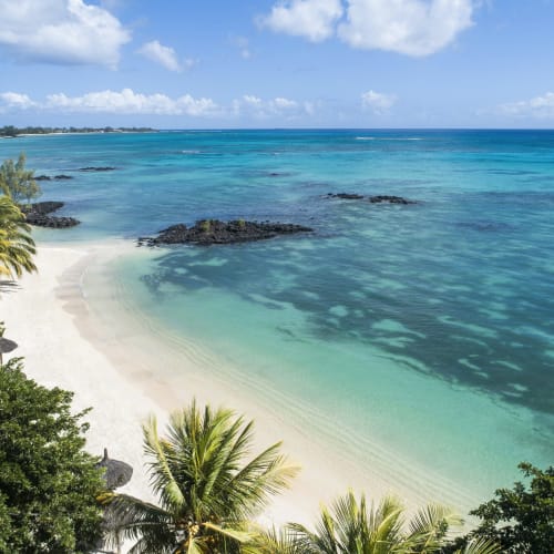 Ein tropischer Strand mit weißem Sand, türkisfarbenem Wasser und üppigem Grün entlang der Küste unter einem bewölkten Himmel.