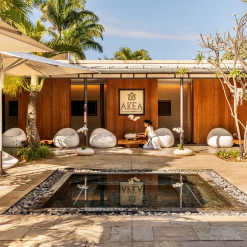 Spa-Bereich mit einem kleinen rechteckigen Becken vor einem Holzgebäude mit dem Schild 'AKEA', umgeben von weißen Liegestühlen und Sonnenschirmen, mit einer knienden Person im Spa-Bereich.