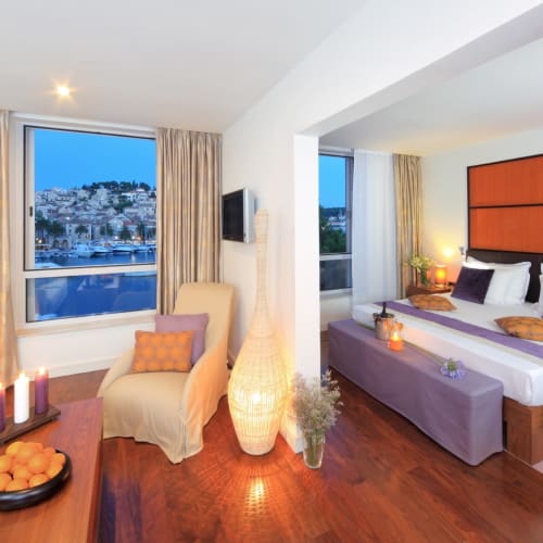 Luxurious hotel suite with a bedroom and a sitting area, decorated with warm lighting and large vases, overlooking a city and water through big windows.