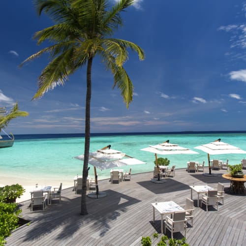 A luxurious beachfront restaurant with white tables and chairs set up on a wooden deck overlooking turquoise sea and white sandy beach, with palm trees and umbrellas under a partly cloudy sky.