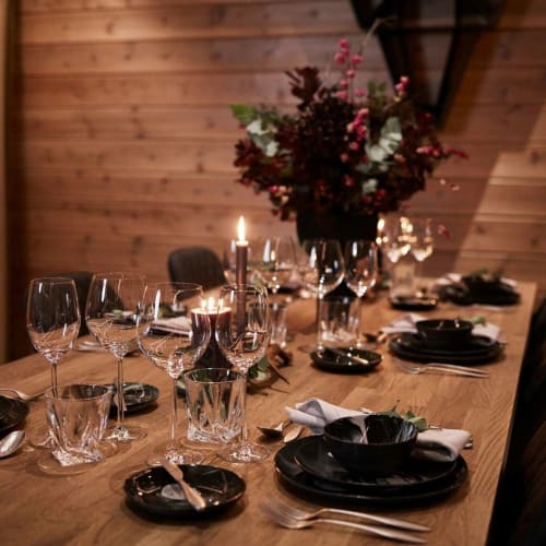 Ein rustikaler Holztisch, der für ein formelles Abendessen gedeckt ist, mit schwarzen Tellern, mehreren Gläsern, Besteck, Kerzen und einem Mittelstück aus dunkelroten Blumen in einer Vase vor einem Holzwandhintergrund.