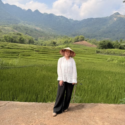 A person stands on a dirt path in front of lush green terraced rice fields with forested mountains and a cloudy sky in the background.