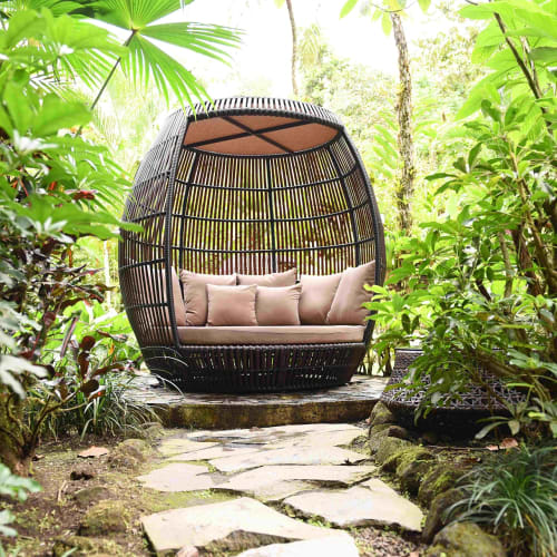 A cozy, outdoor woven cabana with plush cushions surrounded by lush green tropical plants in a garden.