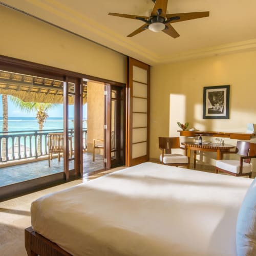 Bright bedroom with a large white bed, wooden furniture, and a ceiling fan, opening to a balcony overlooking palm trees and the ocean.