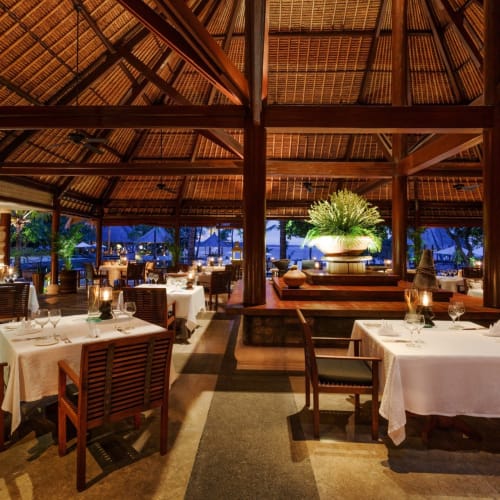 Elegantes Außenrestaurant mit Tischen mit weißen Tischdecken, Holzmöbeln und großen Topfpflanzen unter einem Strohdach.