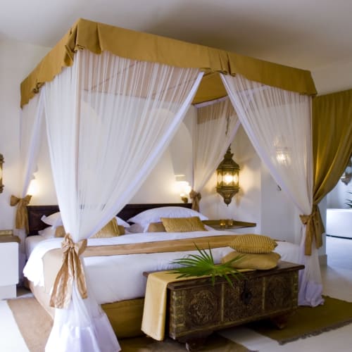 Elegantes Schlafzimmer mit einem vierpfosten Betthimmel in Weiß und Gold, geschmückt mit hängenden Laternen und tropischen Pflanzen.
