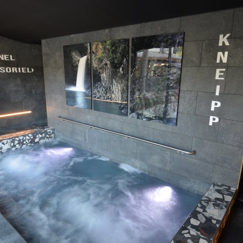 Innen-Whirlpool mit sprudelndem Wasser und einer verschwommenen Person in einem weißen Bademantel, die an einer Bank entlanggeht, mit den Schildern 'Tunnel Sensoriel' und 'Kneipp' an dunklen gefliesten Wänden und einem dreiteiligen Foto eines Wasserfalls über dem Whirlpool.