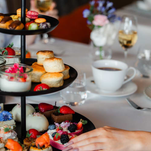 Ein mehrstöckiges schwarzes Serviertablett mit Scones, Macarons, kleinen Desserts und frischen Erdbeeren auf einer weißen Tischdecke. Eine Hand greift zur unteren Etage, im Hintergrund eine Tasse Tee, Weingläser und unscharfe Blumenarrangements.