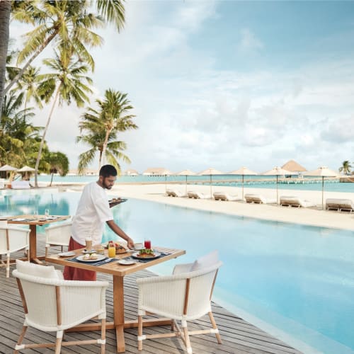 Ein Kellner bereitet Frühstück im Außenbereich am Pool vor, umgeben von Palmen auf einer tropischen Insel.