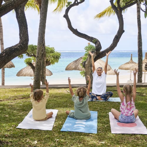 Eine Gruppe von Kindern, die unter Bäumen am Strand auf Matten Yoga üben, während eine Lehrerin ihre Posen beaufsichtigt.