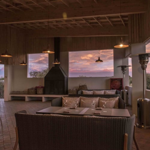 Cozy restaurant interior with large windows offering sunset views, wooden ceiling, and modern outdoor furniture.