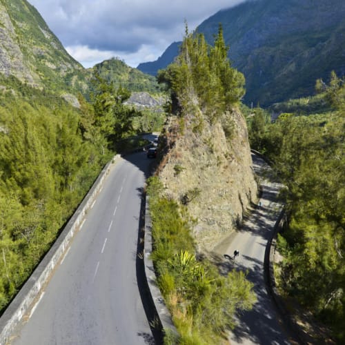 A winding mountain road splits into two narrow paths around a steep rocky outcrop covered with vegetation, surrounded by dense green trees and mountains under a cloudy sky.