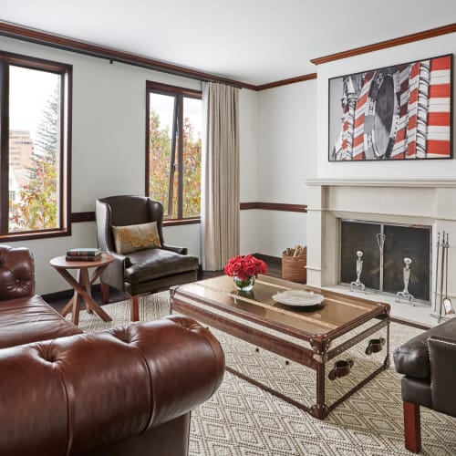 A sophisticated living room with large windows, a leather sofa, armchairs, a fireplace, and an abstract painting, decorated in neutral tones with accents of red.