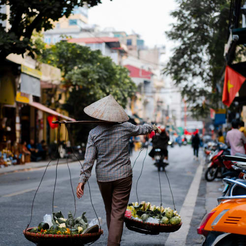 Eine Person mit traditionellem spitzem Hut trägt zwei mit Gemüse gefüllte Körbe an einer Stange über den Schultern auf einer belebten Straße in Hanoi, Vietnam.