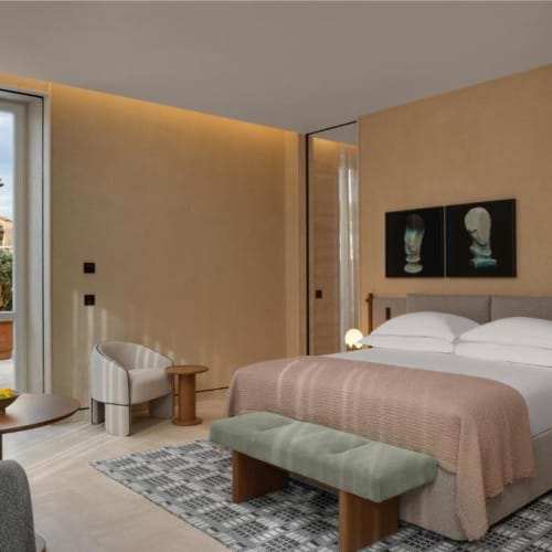 A luxurious hotel bedroom with a large bed, a side table with lamps, a bench, and a sitting area near a glass door leading to a balcony.