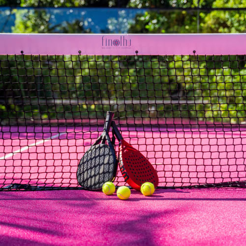 Two padel rackets, one black and one red, leaning against a net on a pink padel tennis court with three yellow balls on the ground in front.
