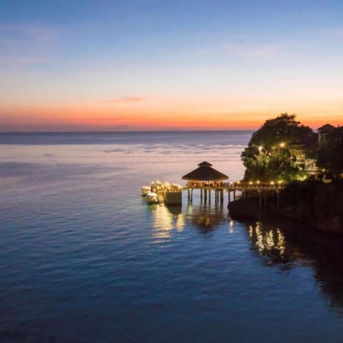 Eine ruhige Flusslandschaft bei Sonnenuntergang mit beleuchtetem Steg und Pavillon, umgeben von Bäumen.