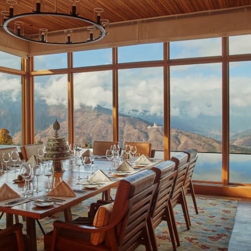 Ein luxuriöses Speisezimmer mit großen Glasfenstern, die Berge und einen See überblicken, für ein formelles Essen mit einem langen Tisch, Servietten, Glaswaren und einem dekorativen Mittelpunkt.