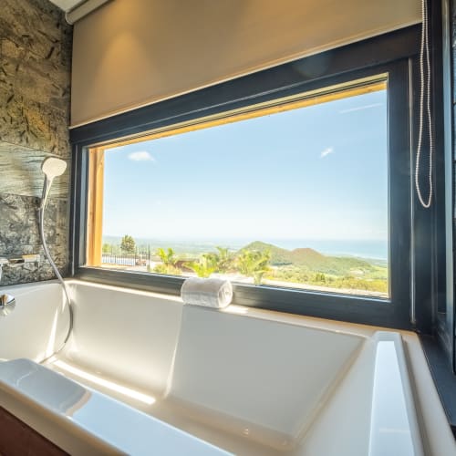 Eine moderne Badewanne neben einem großen Fenster mit Blick auf Hügel, Bäume und das Meer bei klarem blauem Himmel.