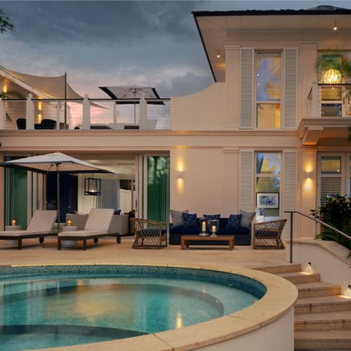 An elegant villa with outdoor seating and a pool, featuring multiple levels and warm lighting, set against a twilight sky.