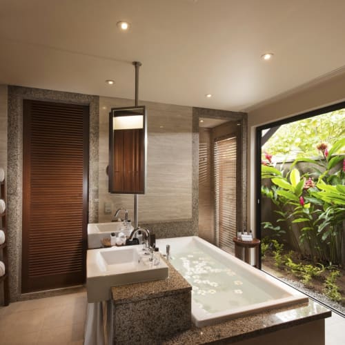 Ein luxuriöses Badezimmer mit einer großen Badewanne, Doppelwaschbecken und Holzelementen, mit Blick auf üppiges Grün durch große Schiebetüren.