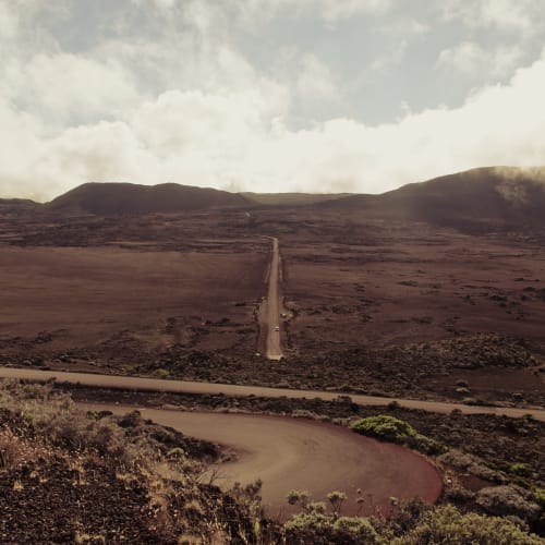 Eine lange, gerade Straße, die durch eine karge vulkanische Landschaft mit zerklüfteten Hügeln im Hintergrund unter einem bewölkten Himmel führt.