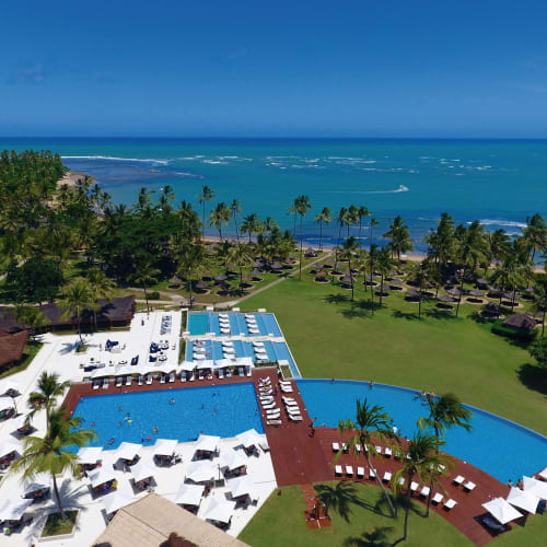 Luftaufnahme eines luxuriösen Resorts mit einem großen Pool, umgeben von Palmen und saftig grünem Rasen, mit Blick auf den Ozean.